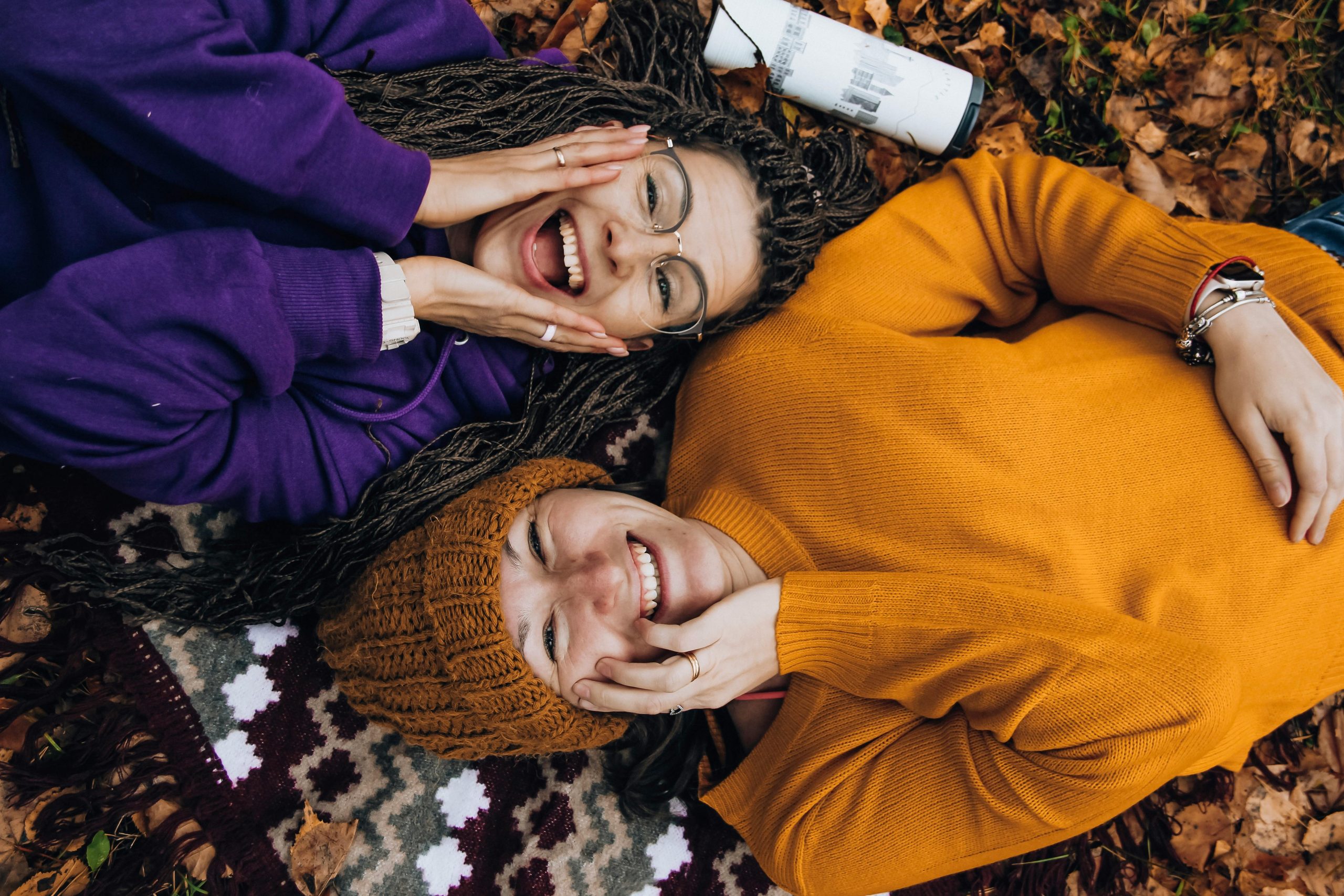 Two friends smiling, enjoying fall colors while lying on autumn leaves outdoors.